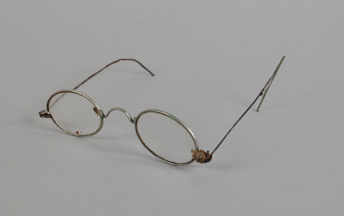 Ovale brilleglass med tynn metallinnfatning. Den ene brillestangen er knekt, den andre har en bue ytterst, og er festet til brillen med tau.