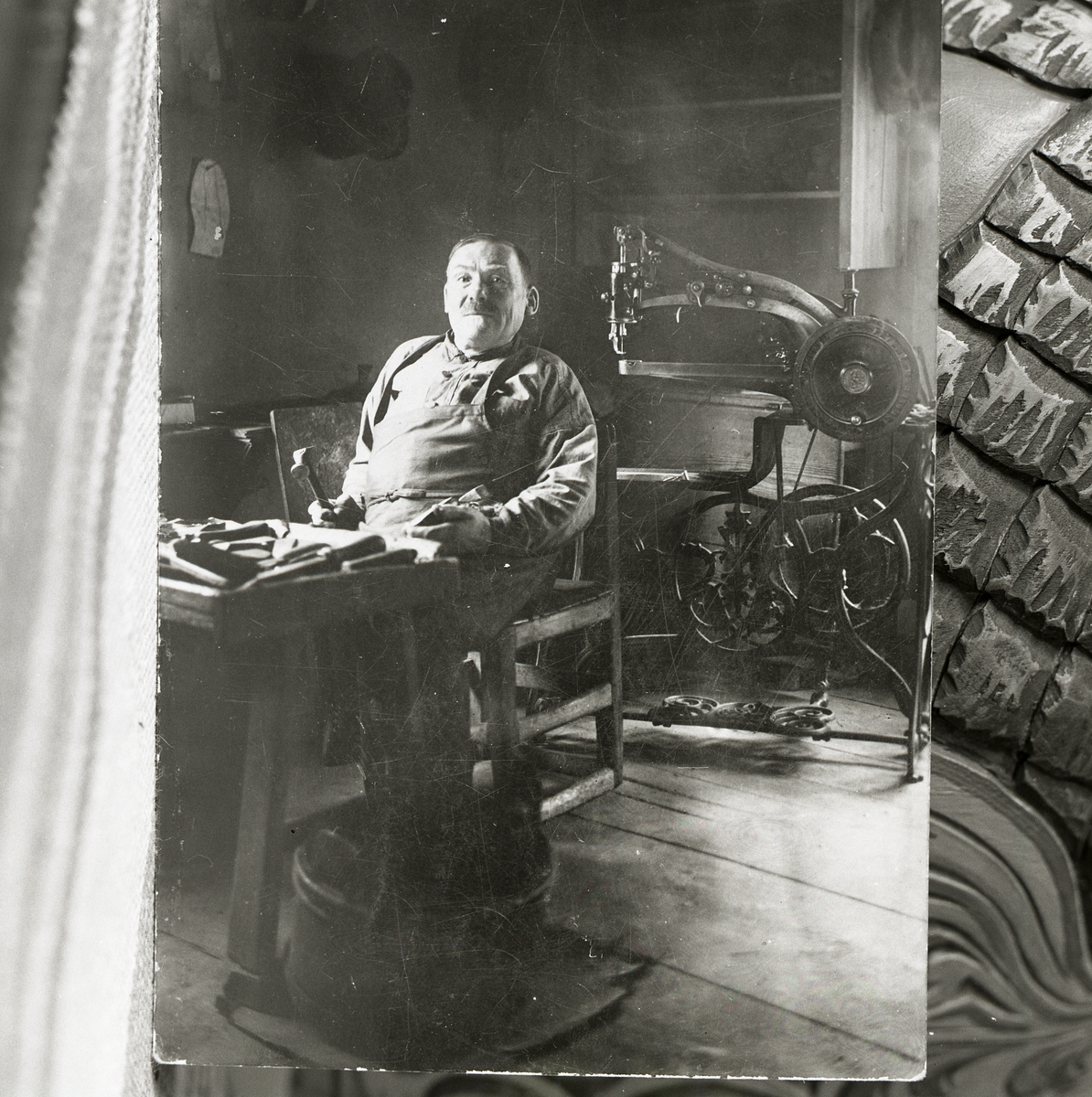 En man med läderförkläde och hammare i handen, sitter i en skomakarverkstad, omgiven av diverse maskiner och verktyg.