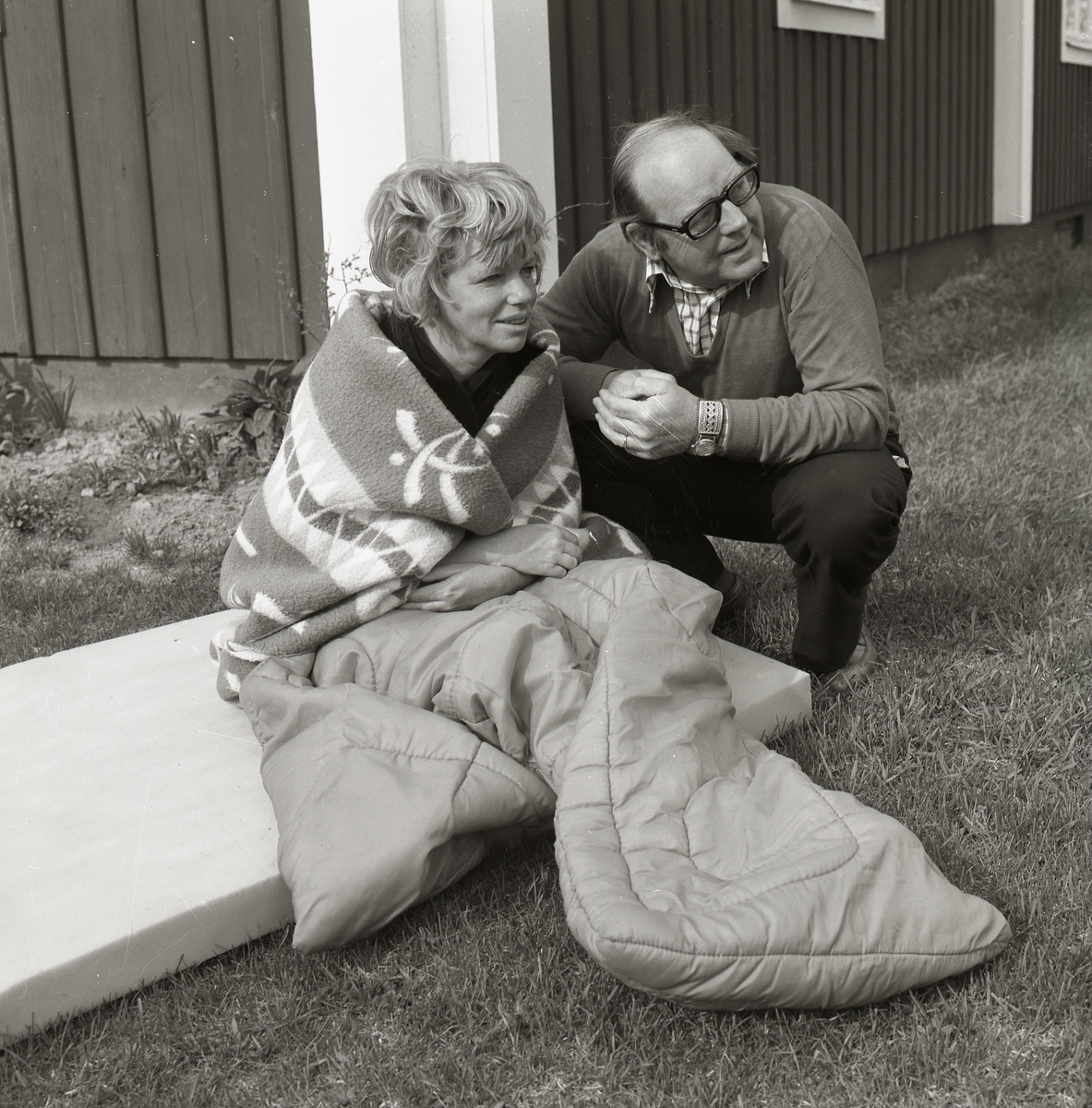 En kvinna sitter på en madrass intill en husknut insvept i en filt och en sovsäck. Bredvid henne står fotografen och filmaren Sven Gillsäter på huk, 29 maj 1976. De spanar efter knipungar och samtalar med varandra.