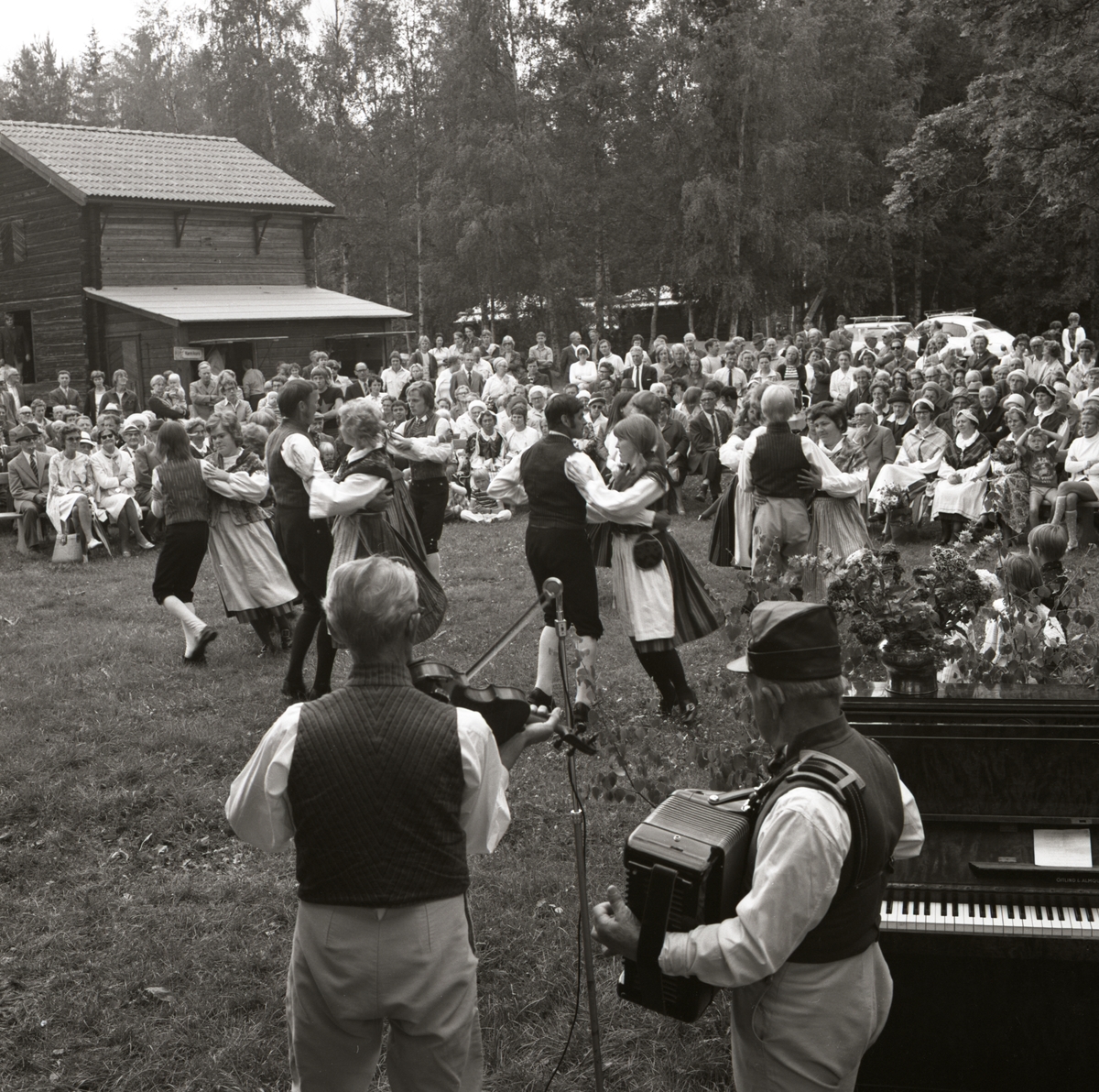 Under ett uppträdande på Rengsjöfesten spelar spelmän dragspel och fiol. Framför dem dansar par iklädda folkdräkt och publiken bakom ser på, 1970 vid Rengsjö hembygdsgård.