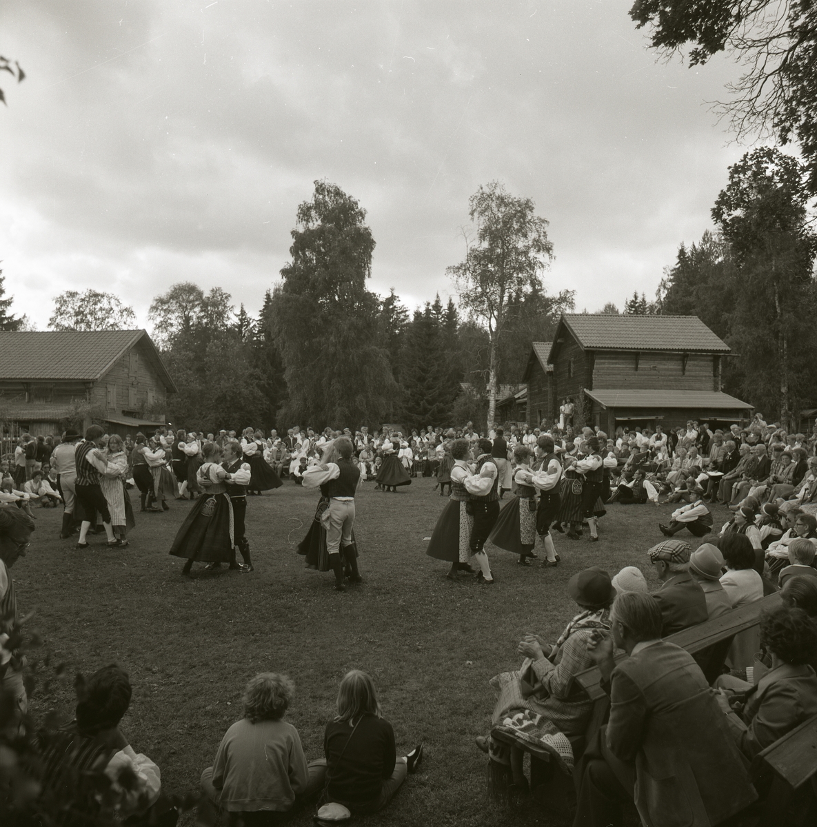 Ett folkdanslag dansar iförda folkdräkter inför en publik. I bakgrunden finns det några härbren, träbyggnad och träd, Rengsjöfesten 1980.
