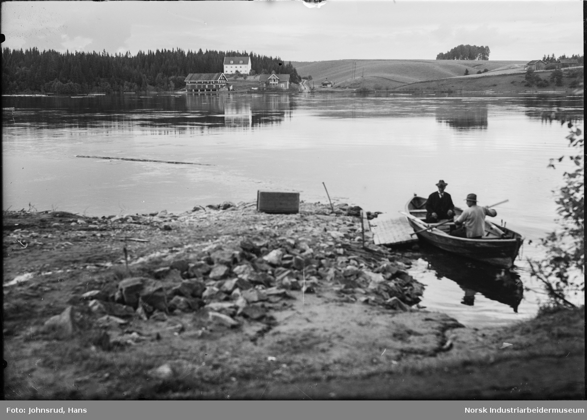 Båt transport over Glomma ved Hagasund - Horgen. To menn sitter i robåt. Gårdsbruk på andre siden av elva.