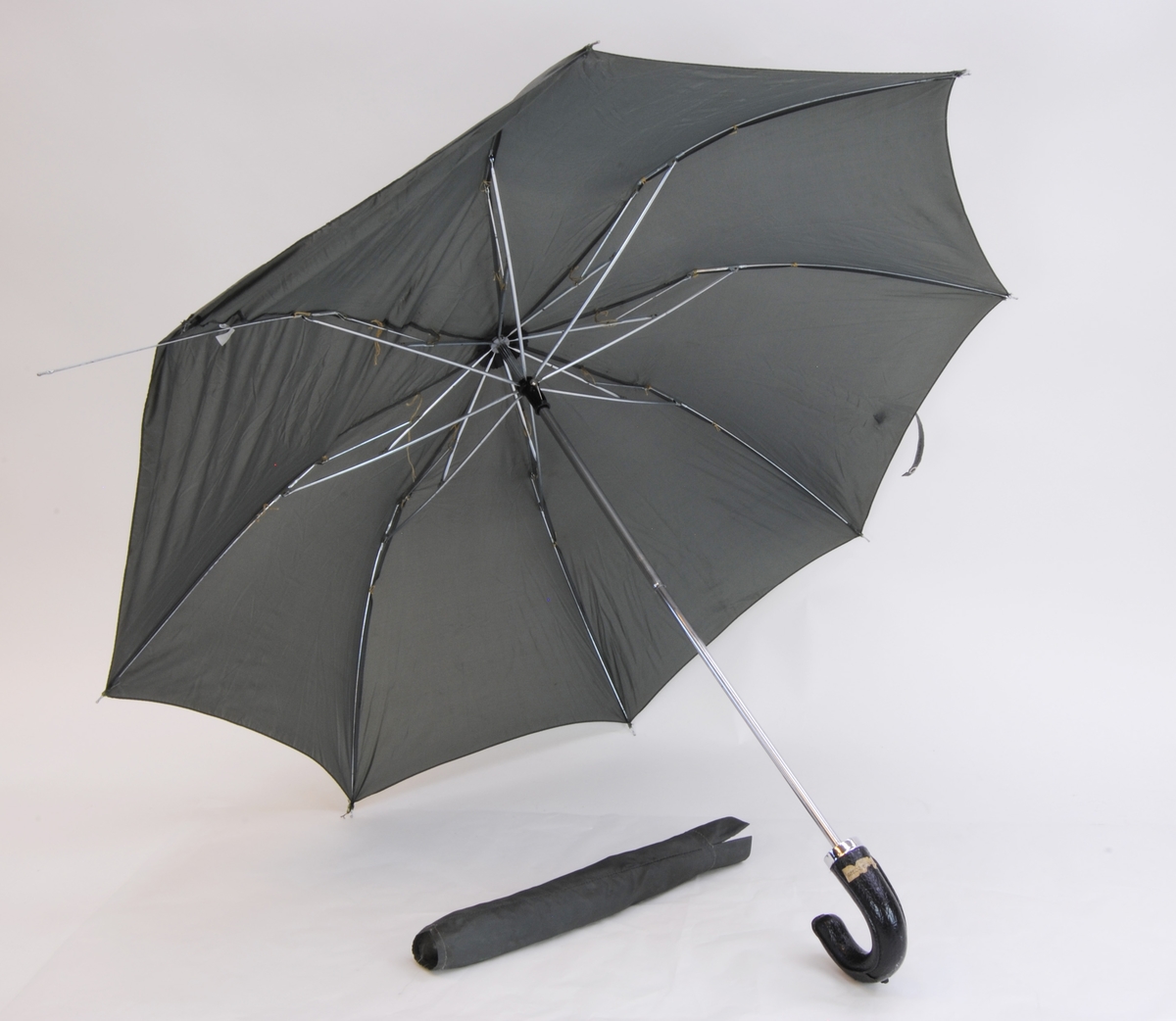 Ett grått paraply med ett böjt handtag av läderimiterad plast. Paraplyt har ett skyddsöverdrag. 
Textilierna är gjorda av 100% nylon.