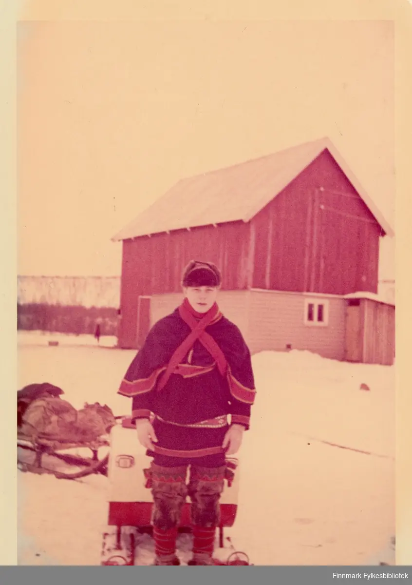 18 år gammel Are Ingvald Hallonen står foran snøscooteren med en kjelke. Fjøs i bakgrunnen. Are Ingvald har på seg luhkka og samisk belte. På føttene har han skaller med skallebånd. Året er 1966.