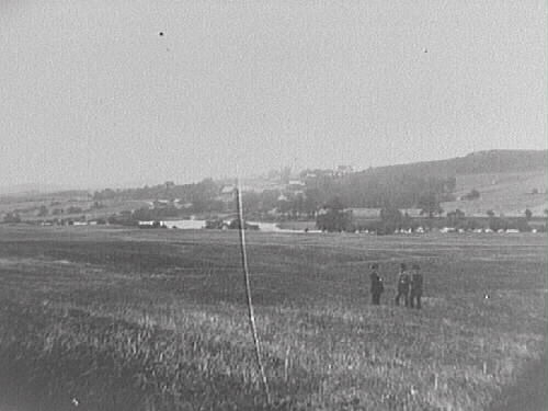 Alfshög socken i Vessigebro. Tre män står på ett fält med ån och kyrkåsen i bakgrunden.