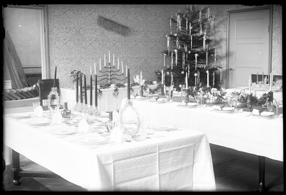 Exempel på juldukningar visas på Husmodersföreningens julmässa. Fyra bord med linnedukar och porslin står uppställda och i rummets ena hörn står en julgran.