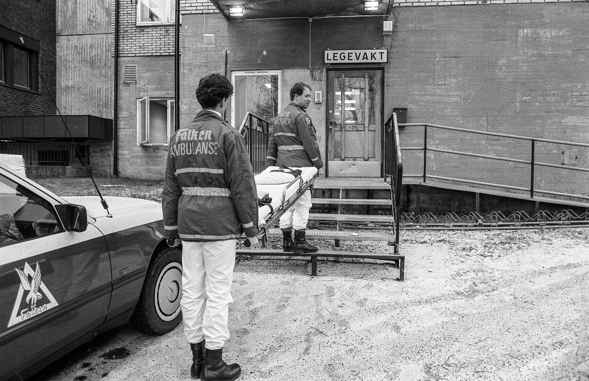 Legevakten i Ski / Oppegård ved Ski sykehus. Ambulansepersonell, Roy Pedersen og Frank Rødsand, fortviler, står med båre mellom seg utenfor på en jerntrapp.