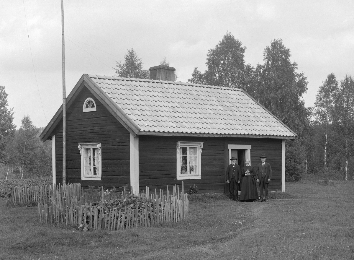 Den prydliga backstugan Bäckhult på Klockarps ägor i Svinhult. Framför stugan ses makarna Karl Ludvig Jonsson och Charlotta Pettersson. Mannen till höger är möjligtvis parets yngste son, Gustaf Adolf.