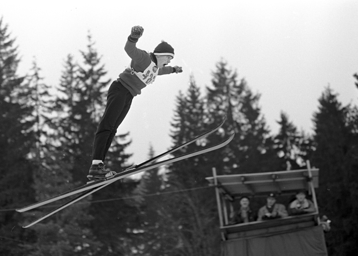 En skihopper i svevet, dommertårn i bakgrunn, Bekkelaget skirenn i Bekkelagsbakken. Fotografert 8. mars 1964.