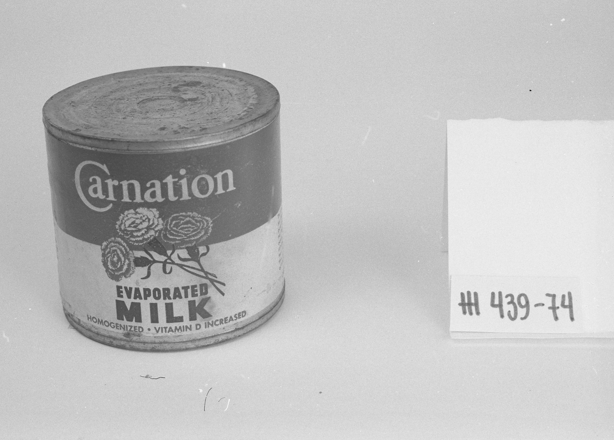Boks blikk, falset, loddet, sylinderform. Etikett papir hvit/rød. Mrk. "Carnation", "EVAPORATED MILK", "HOMOGENIZED", "VITAMIN D INCREASED". Tre nelliker varemerke. Rusten. Uåpnet, inneholder kondensert melk.