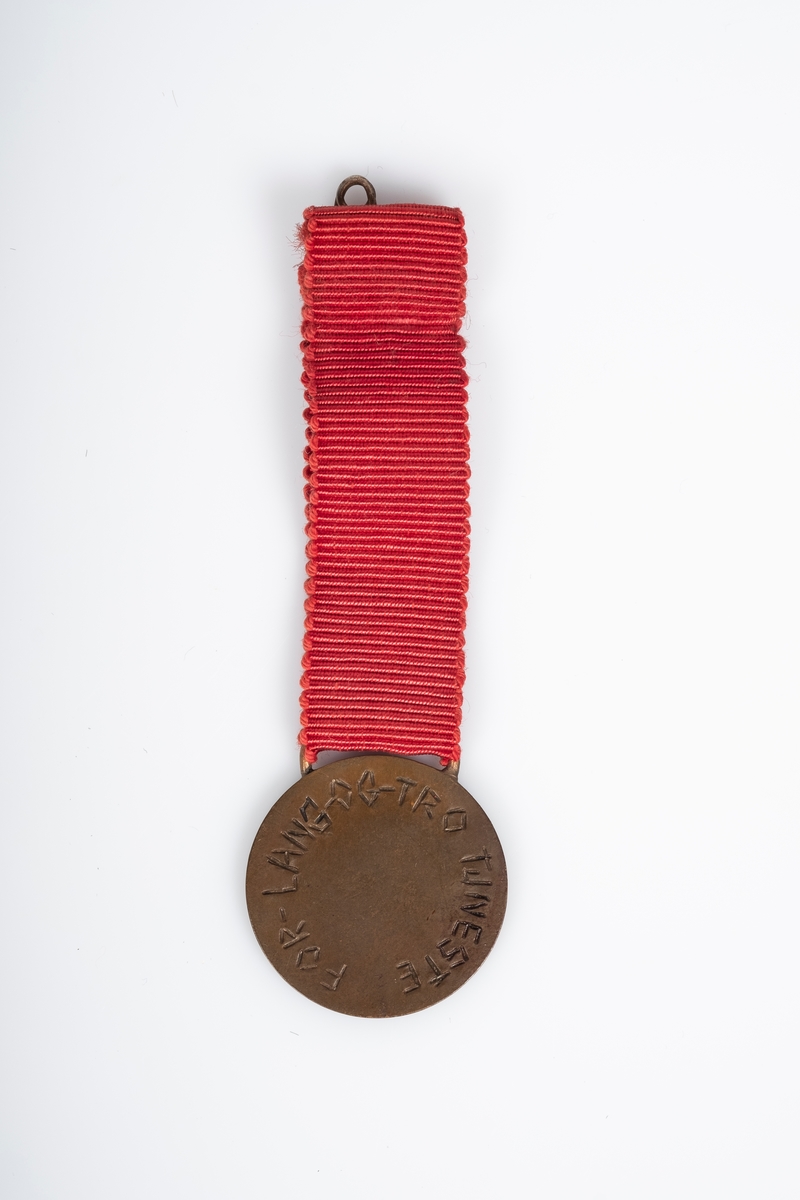 En rund medalje i metall med rødt bånd. På medaljens bånd er det påfestet en sikkerhetsnål. Medaljen har H7-symbolet. På baksiden av medaljen er det tekst som ser ut til å være inngravert på hånd.