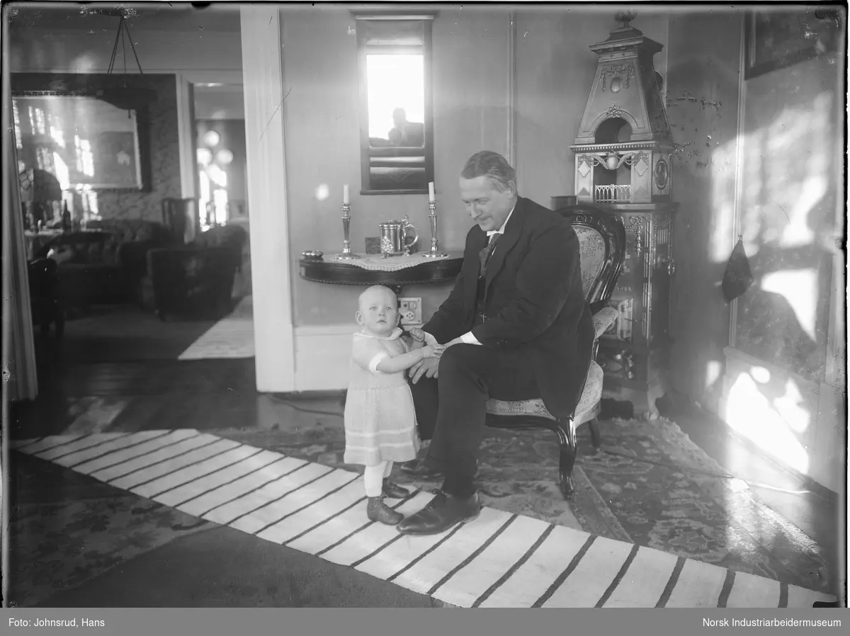 Mann holder barn som står foran han i hendene. Fotografen synlig i speilet i bakgrunnen.