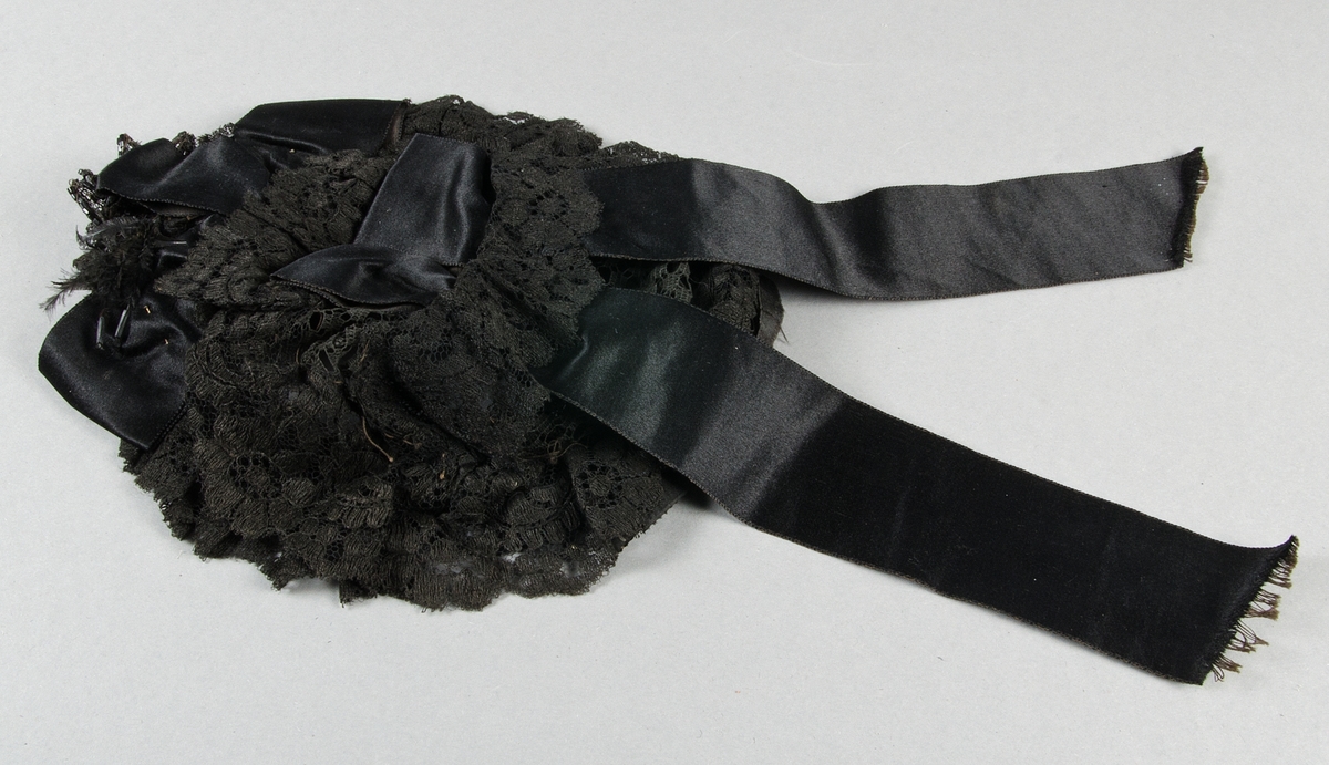 Hårklädsel av svart bomullspets med svarta duchesseband på oval ståltrådsstomme. Prydd med svarta pärlbroderier och fjädrar.

