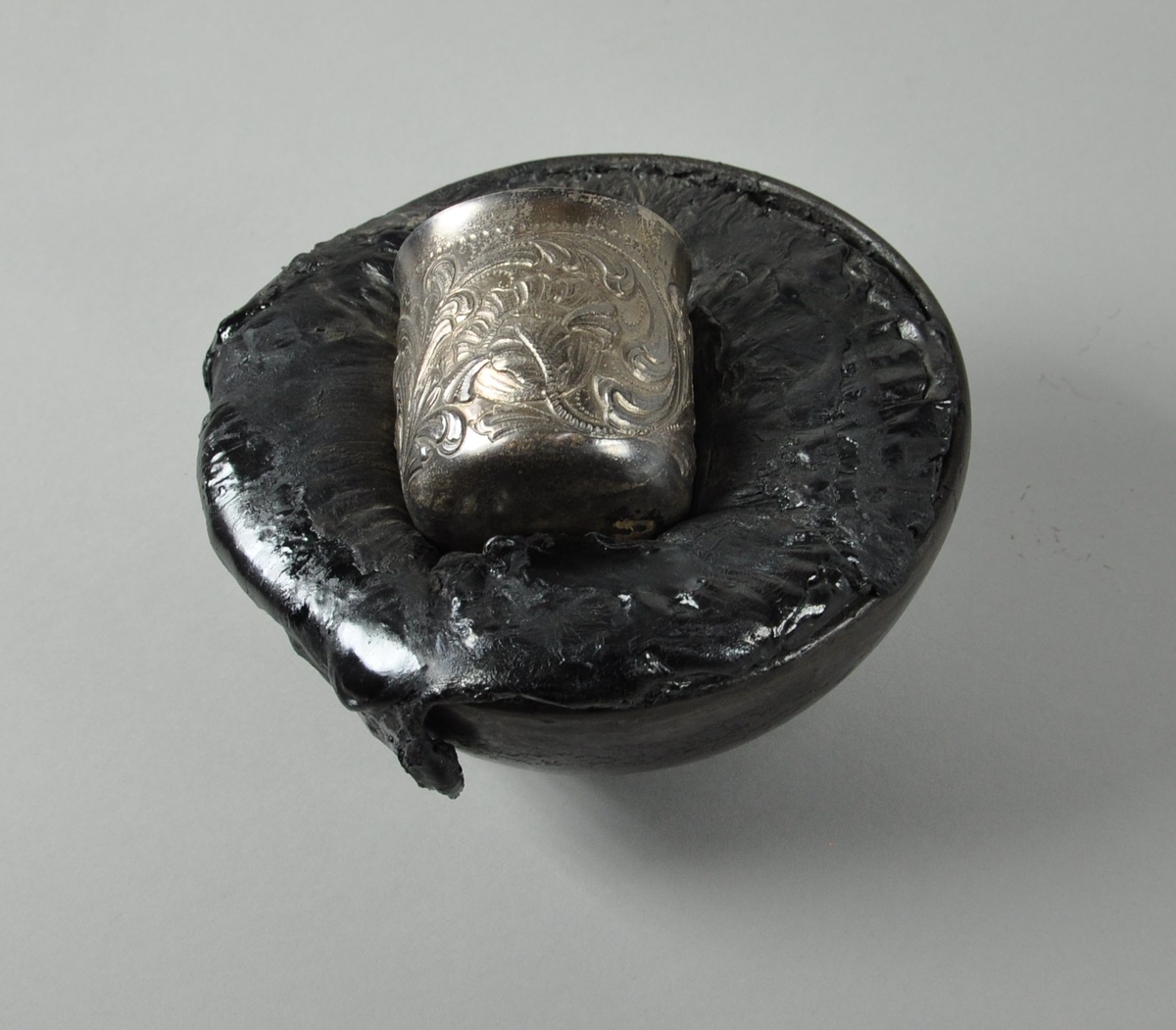 Rund bolle av metall med stivnet bek oppi. I beket ligger et krus av sølv med dekorativ siselering. Kulen står på et sammenfoldet belte av skinn.
