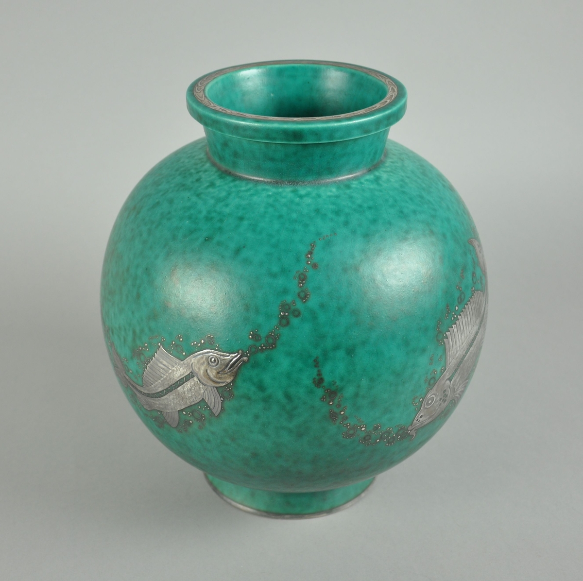Grønn vase av keramikk. Vasen har en rund form med sokkel og rand. På oversiden av randen går det en dekorborde. På korpus er det gravert dekor av metall, forestiller fisk som blåser luftbobler.
