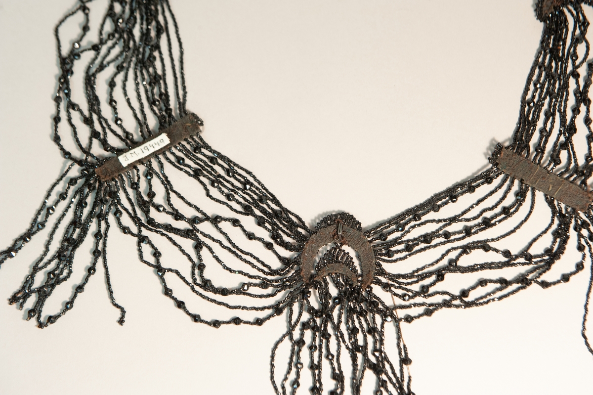 Ett klänningsgarnityr med pärlrader av stenkolspärlor uppträdda på silkestråd. Tre hakar finns på baksidan för att kunna fästa pärlraderna i plagget.