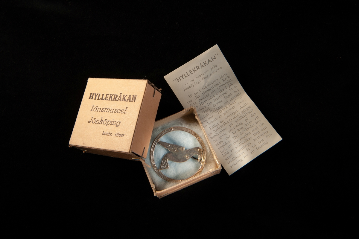 En silverbrosch i form av en ring med en avbildad spillkråka inom. Med broschen följer en tillhörande ask samt en informationslapp.