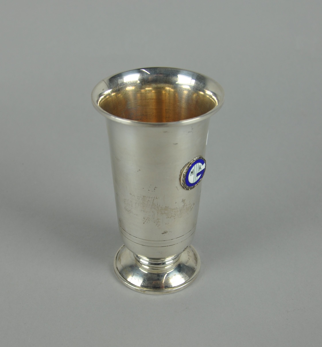 Pokal av sølv. Pokalen har sylindrisk form med utvidelse mot toppen. Pokalen har sokkel, og på korpus er det et emblem med laurbærkrans og en blå G.