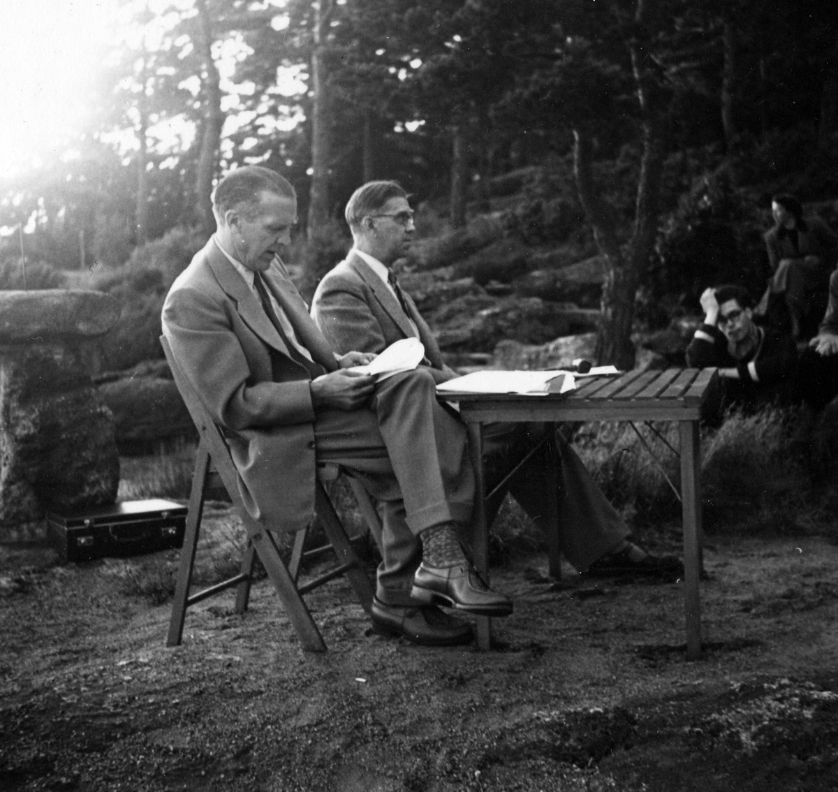 Två män sitter vid ett bord utomhus på ett årsmöte. I bakgrunden syns mötesdeltagare. Anteckning på kortets baksida "Årsmöte 1954".