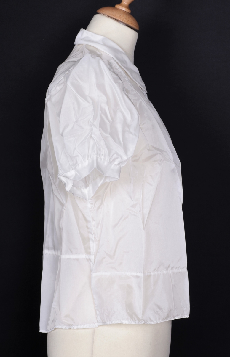 Hvit bluse sydd av fallskjermsilke (nylon). Fallskjermen blusen ble sydd av ble droppet på Vindflomyrene 28. februar 1945, og ble tatt i mot av Harald Alm. Harald Alms kone, Paula Windland, fikk blusen sydd av sin tante, Maria Skogstad.