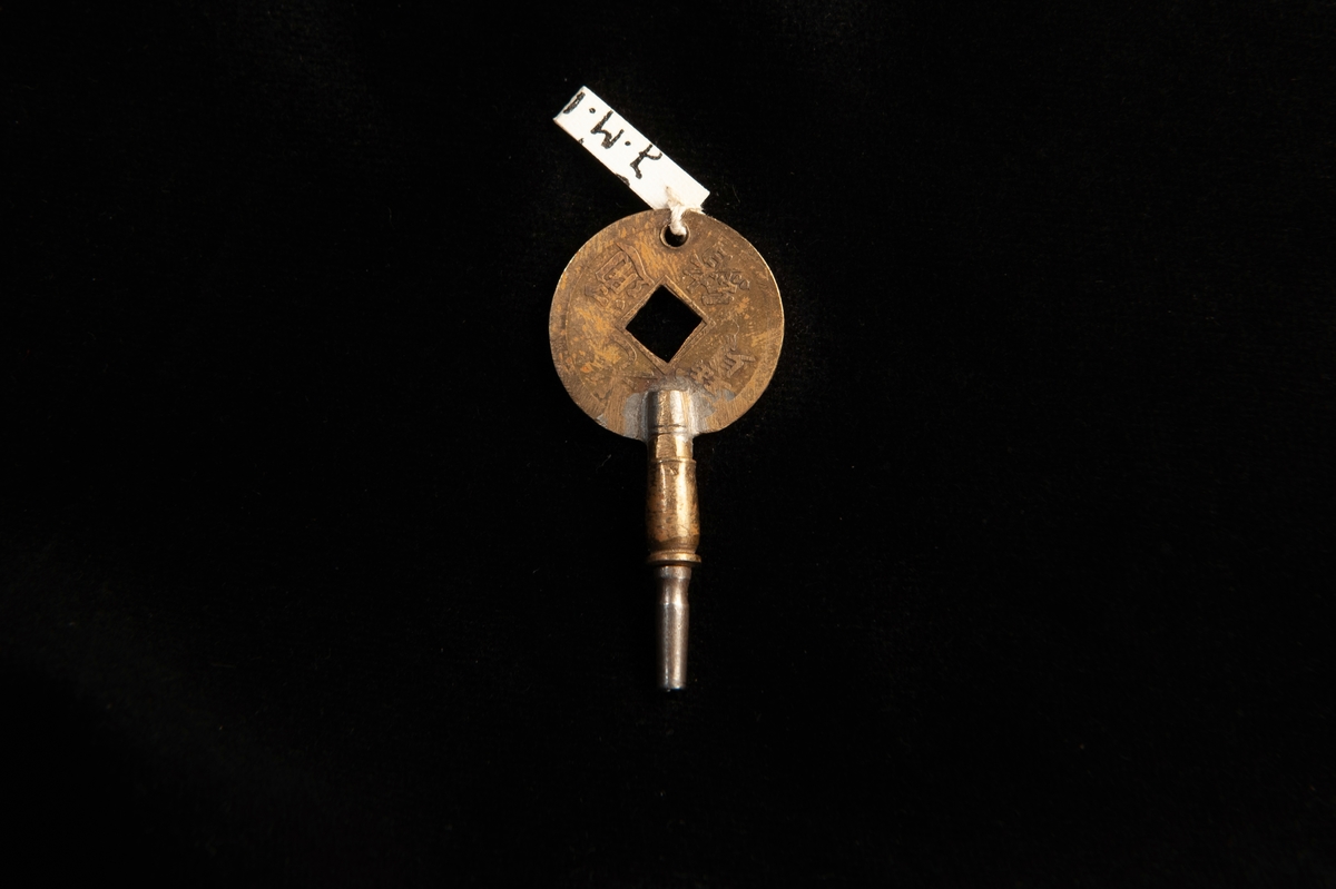 En urnyckel av mässing med runt handtag, inuti handtaget är ett snedställt kvadratiskt hål. Uppdragningsdelen är av metall. Det runda handtaget är gjort av en kinesisk penning.