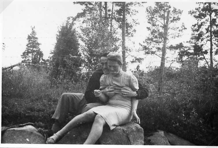En man och en kvinna sitter i naturen på en sten. Han omfamnar henne hårt och greppar hennes bröst.