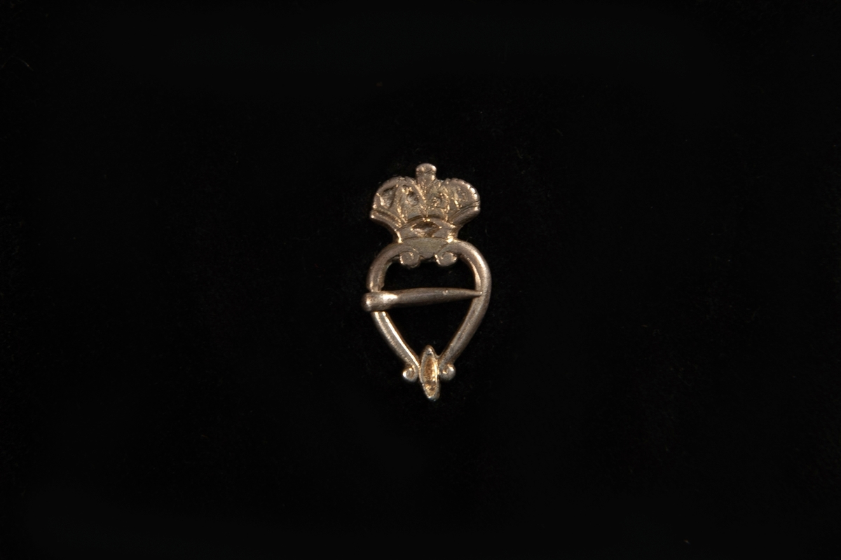 En hjärtformad sölja av graverat silver, med krona överst. Saknar stämplar. Påminner starkt om en s.k. bröstsölja.