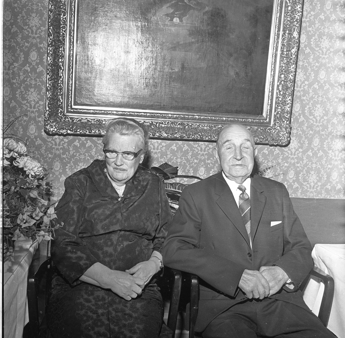Alma och Josef Larsson i Uppgränna Bultagård firar guldbröllop, 1967. De sitter i varsin stol framför mönstrad tapet med stor oljemålning. Till vänster ett bord där en blombukett skymtar.