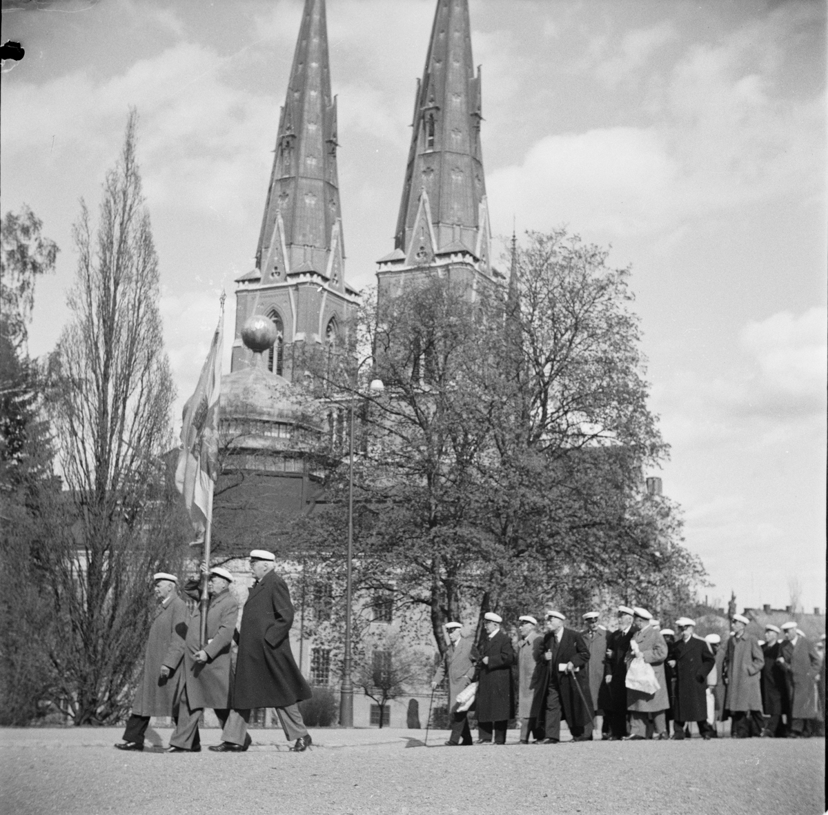 Flera personer iförda studentmössor tågar tillsammans i Universitetsparken, Östgöta nations minneslandskap, Uppsala 1949
