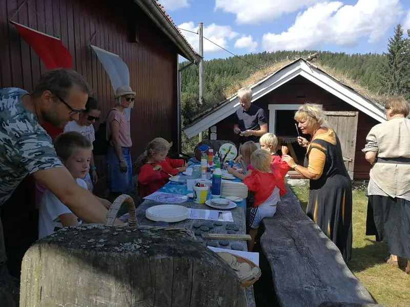 Skjoldverksted under Middelalderdagen på Almenninga. Store og små er samlet rundt det lange bordet på baksida av Vestun. Vestun er malt rød. Unger er i full gang med å male skjold (papptallerkener med gummistrikk på).