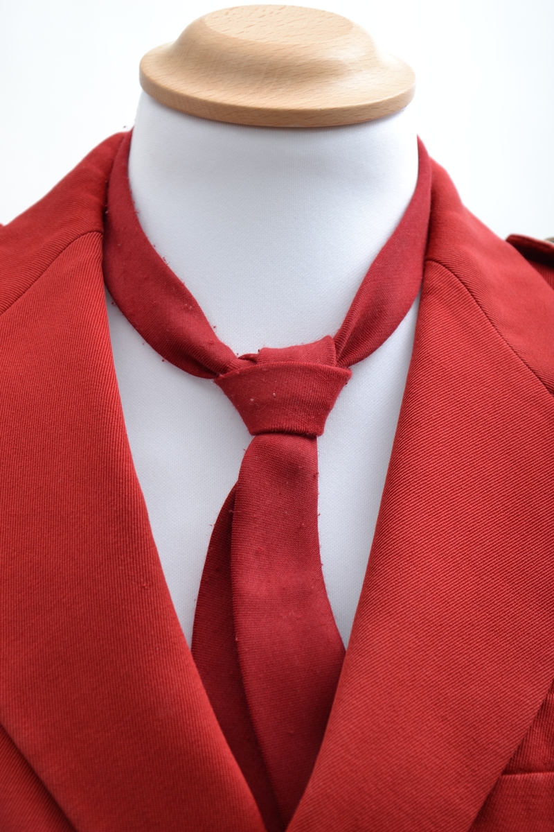 Korpsuniform bestående av jakke, skjorte, slips,to skjørt, hansker og lue. Jakken er dobbeltspent med 4 metallknapper, skulderklaffer og et felt med et smalt hvitt bånd på armene. Skjørtene er grå og plisserte og lukkes med knapp og trykknapper. Enkel hvit skjorte med rødt slips til. Uniformsluen er rød med hvite bånd og dusk i pannen. Et par hvite bomullshansker hører til. Jakken er i størrelse 10 år.