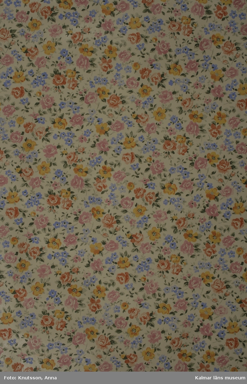 KLM 39827. Tapet i papper, 1 st rulle. Oblekt pappersfärgad bakgrund med blomstermotiv av små blå och gula blommor med gröna blad samt rosa och röda rosor. Mönstret är litet och tätt. Datering: 1940-1950-tal.