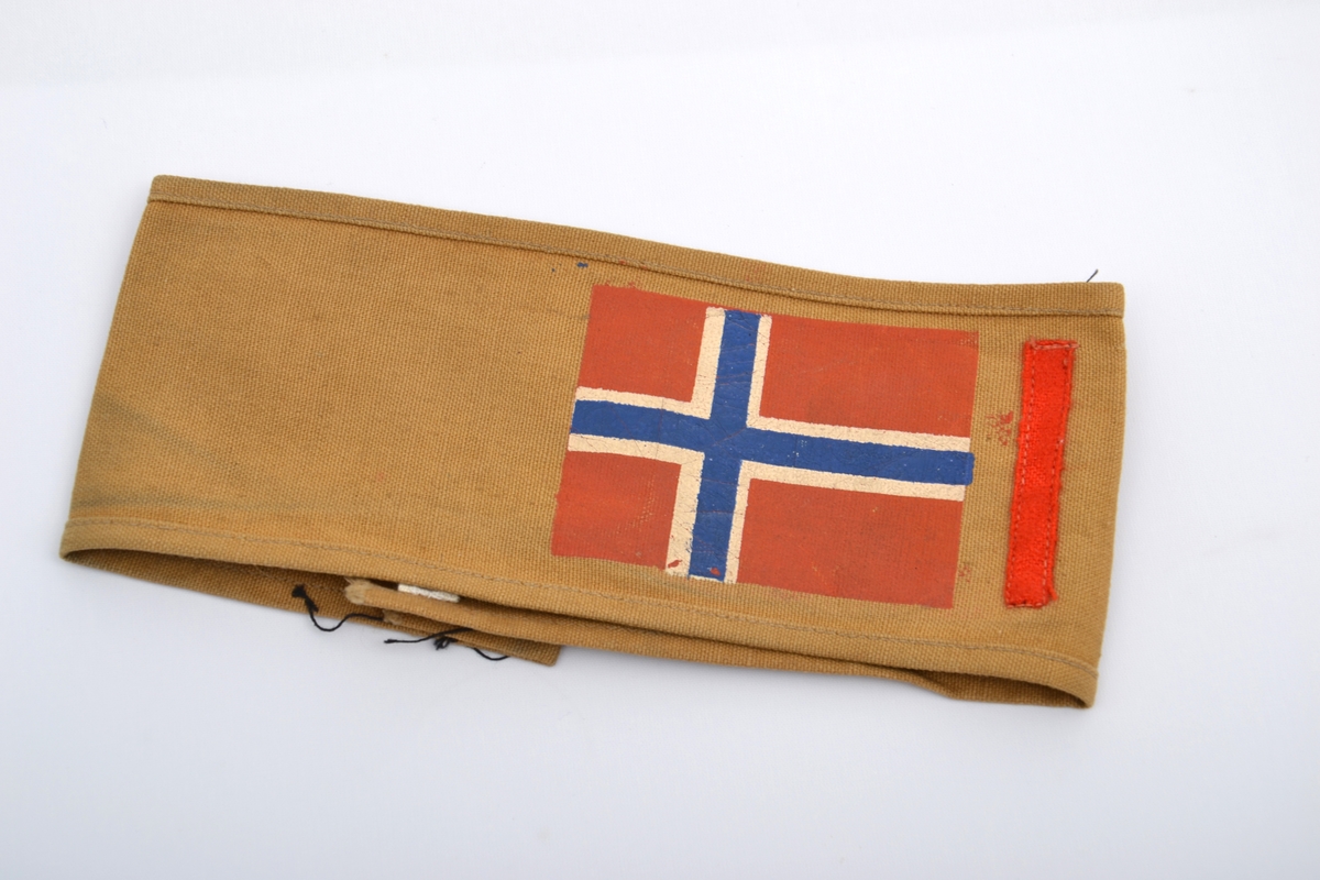 Armbind framstilt i brunt, grovt lerretsstoff med det norske flagg og en smal stripe.