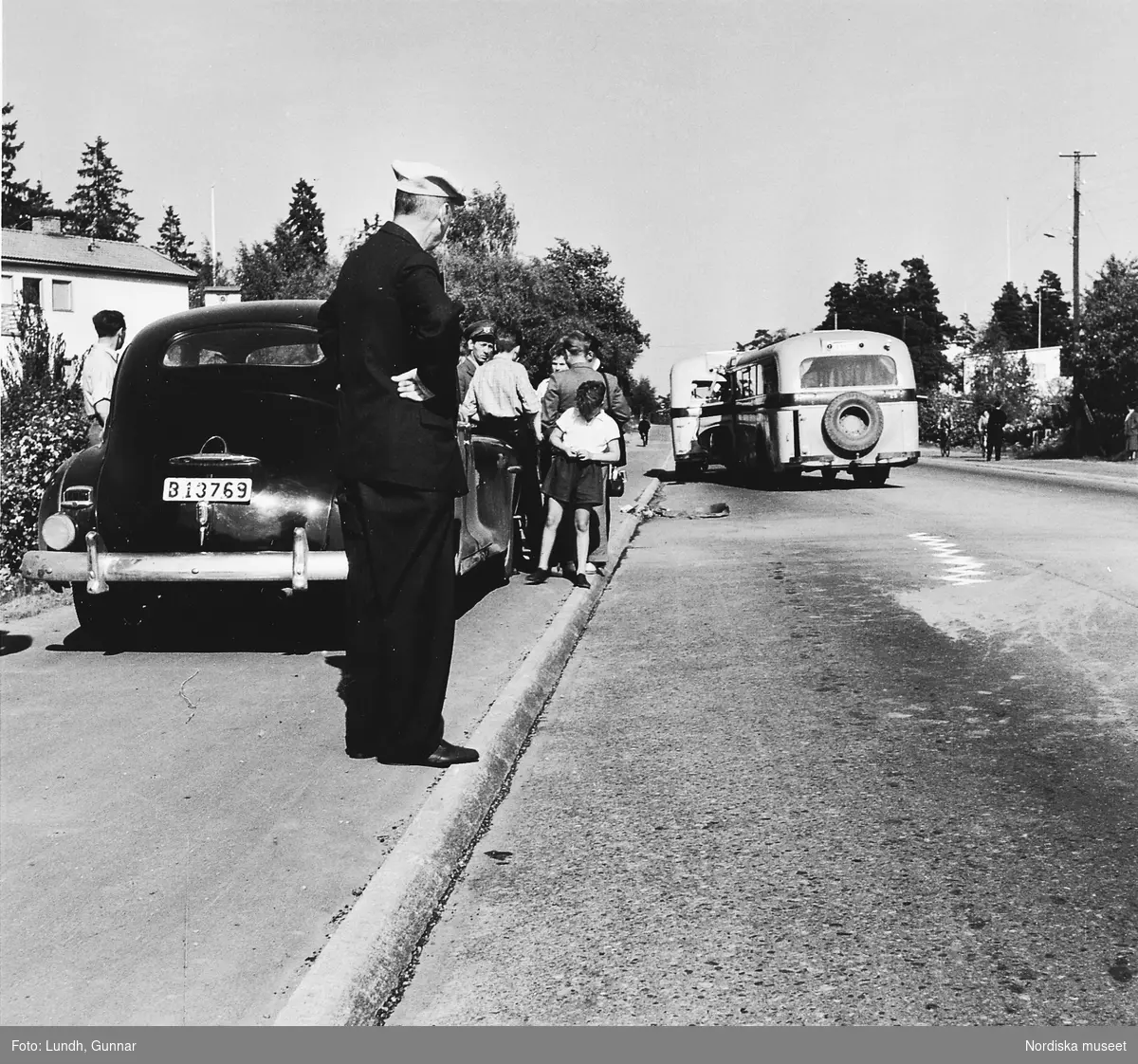 Två bussar har krockat på gamla Värmdövägen, Stockholm, år 1950. En man står på trottoarkanten och tittar, bakom honom en grupp människor vid en bil.