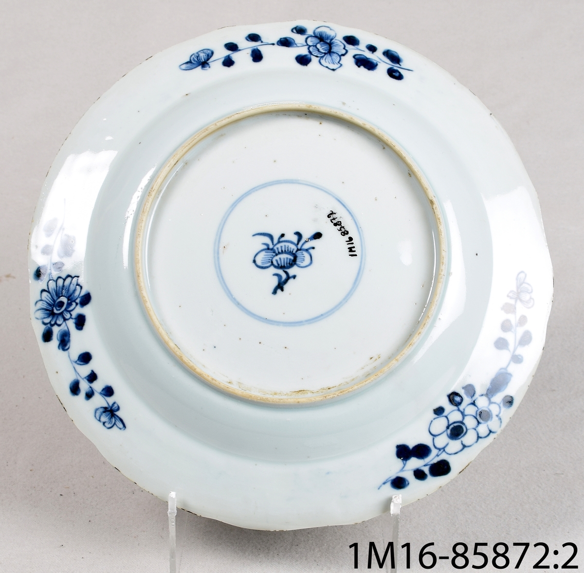 Tallrik av vitt porslin med svagt vågig kant. Har handmålad blå dekor föreställande blommor och insekter. Har dekor både på ovansida och undersida.
