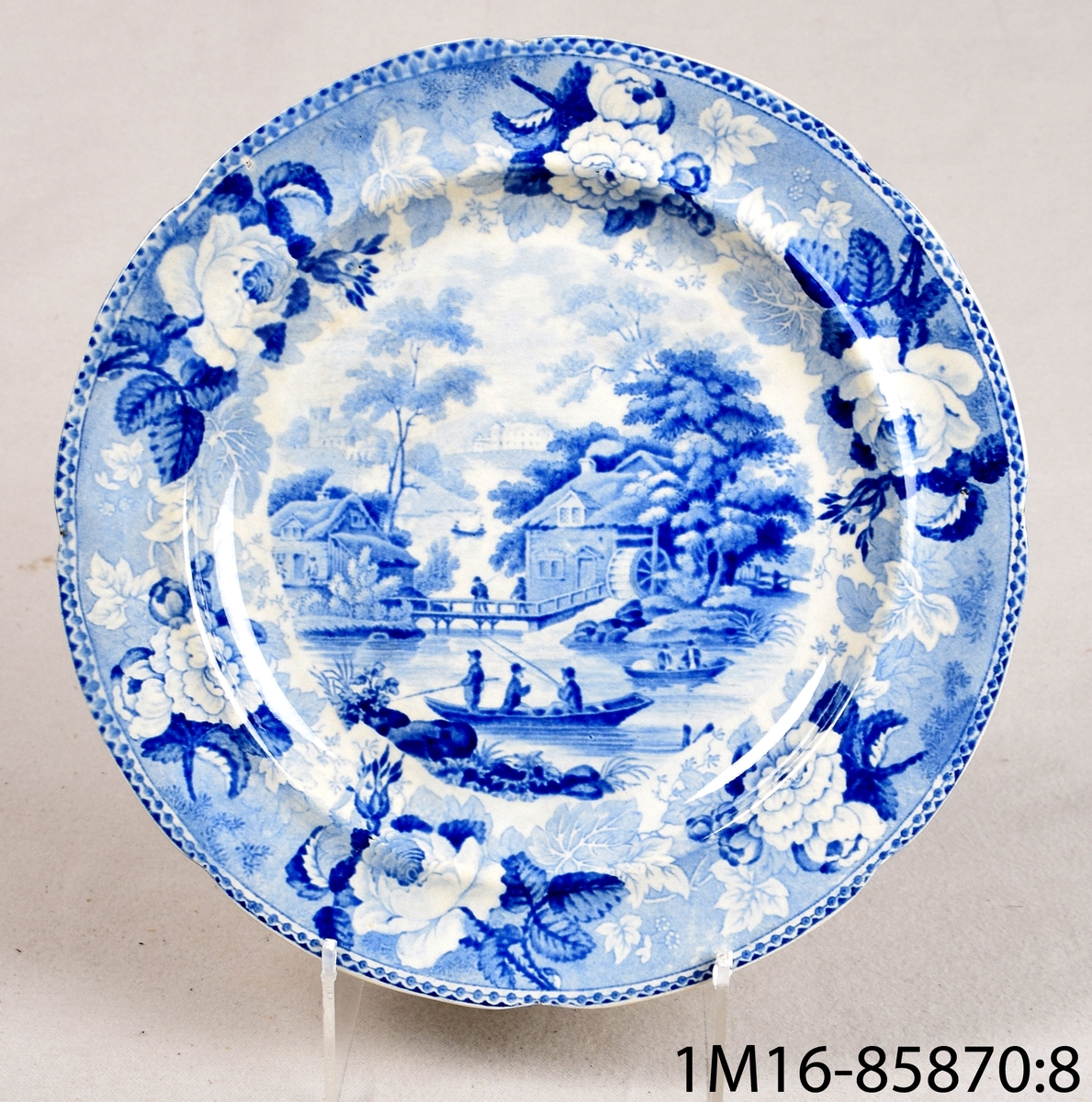 Vit rund tallrik av flintgods med tryckt blå dekor. Dekornamn: 1828 års landskapsmönster. Tillverkare: Rörstrand