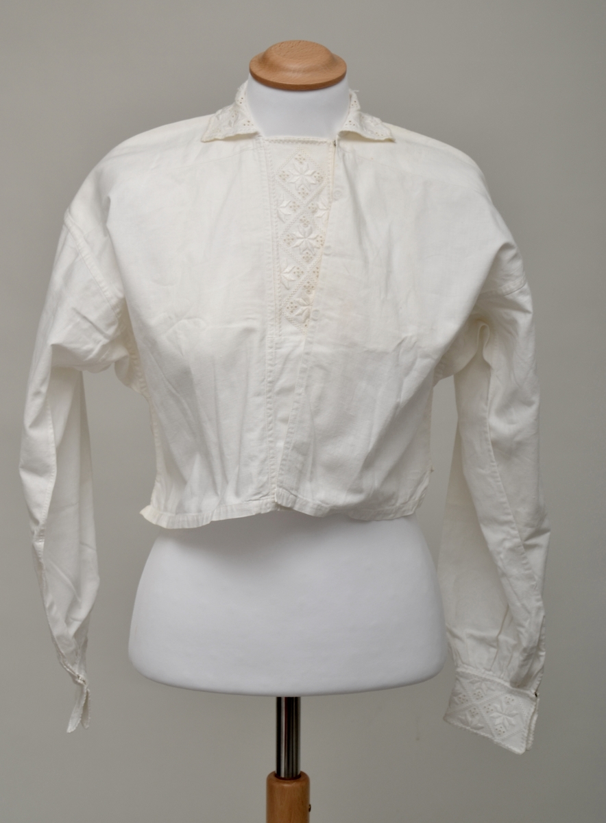 Hvit bomullsskjorte med innfelte stykker med hardangersøm på sultanstoff i stolpe, mansjetter og krage. Lukkes med fire trykknapper i front (to mangler) og en trykkanpp på hver mansjett.