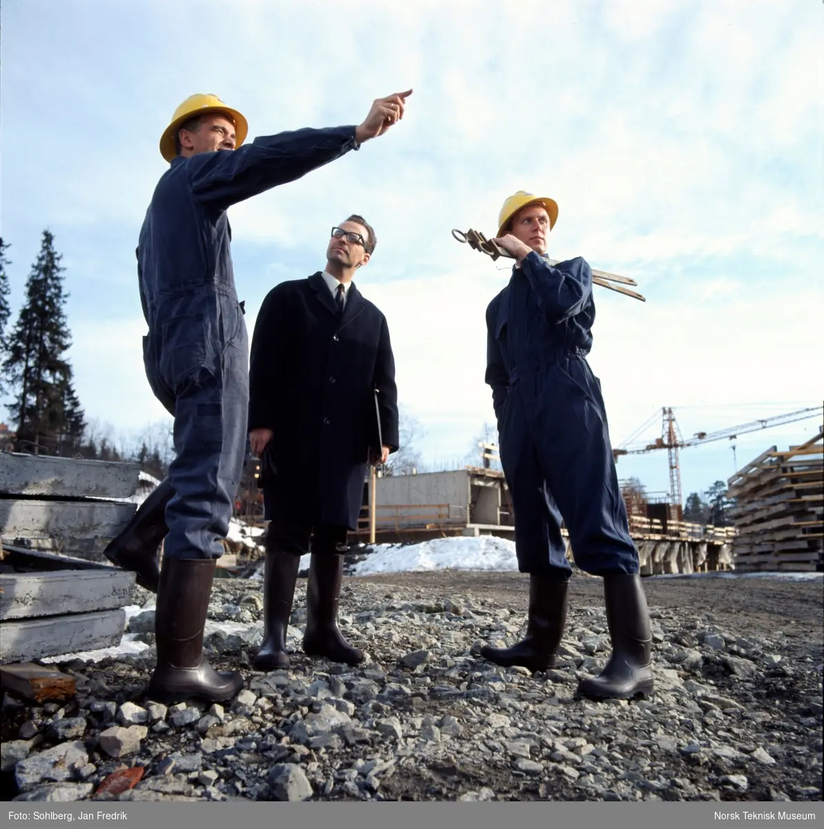 Reklamefotografi for Viking gummistøvler. Tre menn på en byggeplass, to iført kjeledress, hjelm og støvler, den tredje iført frakk og støvler.