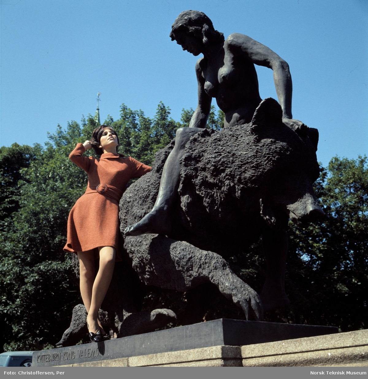 Motebilde i farger. Kvinnelig modell i rødbrun ullkjole av norsk fabrikat, fotografert foran skulpturen "Kvitebjørn kong Valemon" (1933-36) av Dyre Vaa på Ankerbrua/ Eventyrbrua i Oslo, 1968. Bildet er en del av en serie der ett av dem er publisert i ukebladet Det Nye nr. 39, 1968.