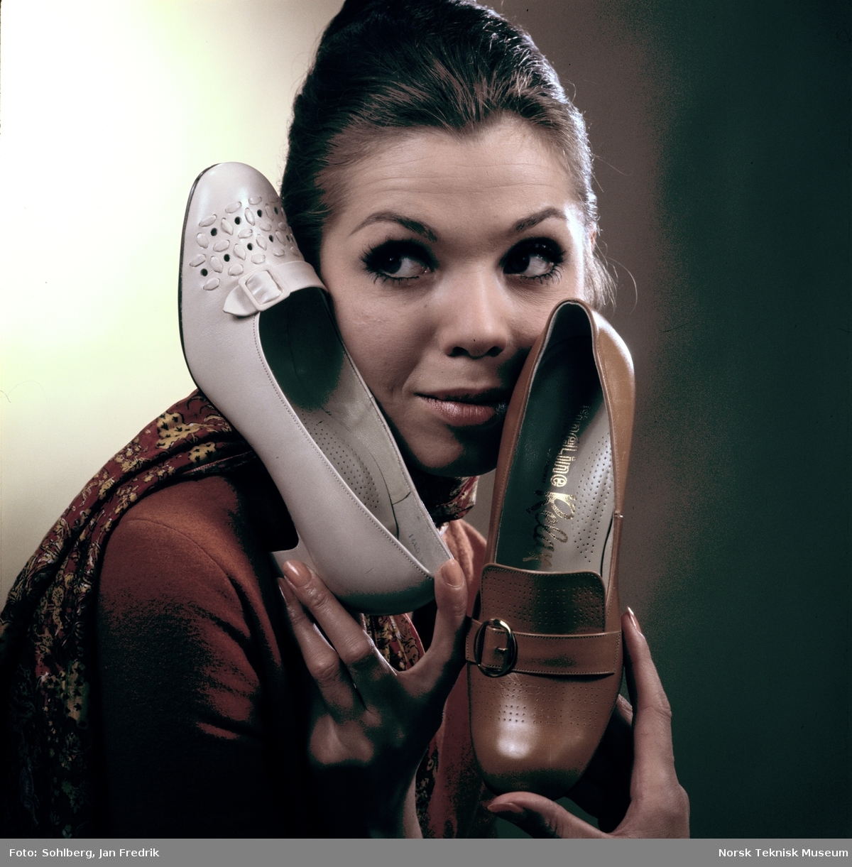 Tidlig norsk reklame- / motefoto. En kvinnelig modell holder opp en brun sko med hæl og spenne, hun virker fornøyd med tidens skomote. Hun kikker henført på skoen.