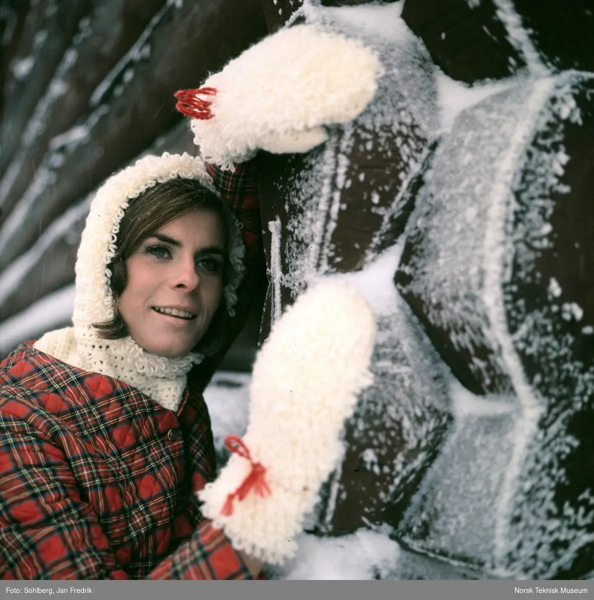 Kvinnelig modell viser vintermote. Hun har en hvit heklet genser med hette, laget av tykt, loddent garn under en skotsk-rutete vattert jakke. Store votter av samme type garn som hettegenseren. Hun lener seg mot en vegg av laftet tømmer.