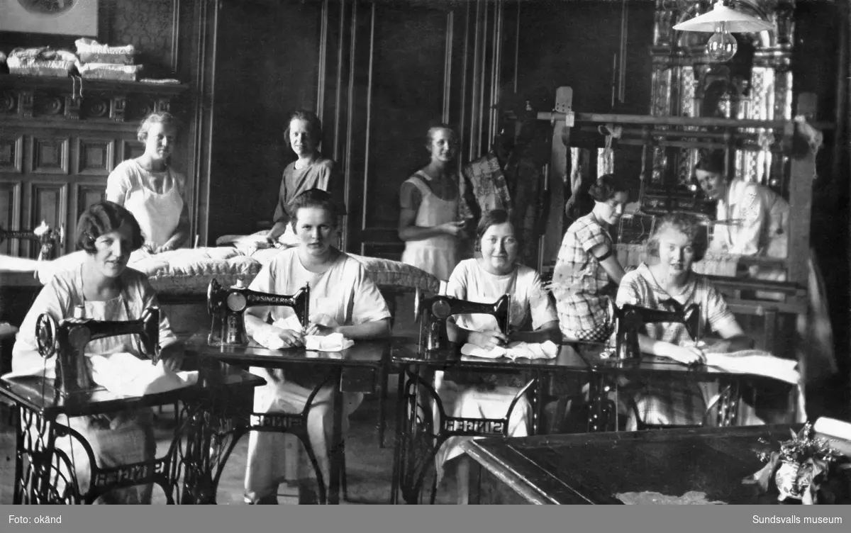 Gruppbild med elever vid symaskiner vid Vita Bandets husmodersskola. "Slöjdgruppen". Vårterminen 1926. Vita Bandet huserade vid denna tidpunkt i en lägenhet i Thuressonska huset vid Nytorget. Först 1928 införskaffades en egen fastighet på Tjärngatan 4.