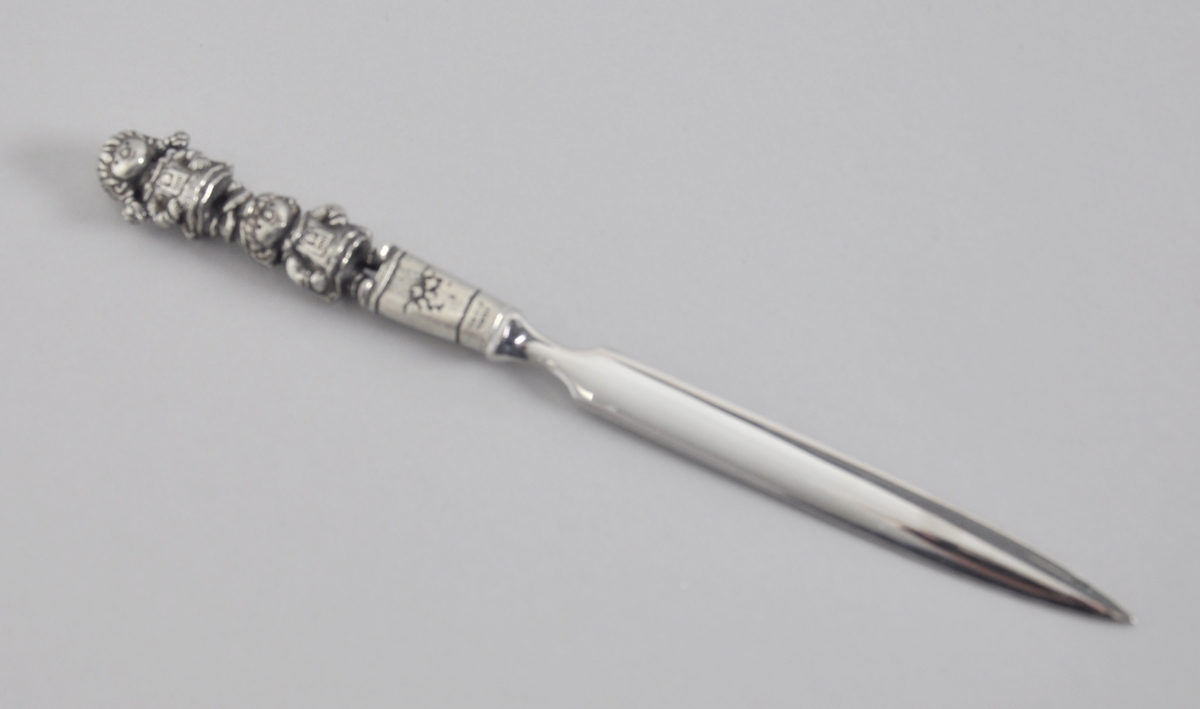 Brevkniv av tinn med maskotene Kristin og Håkon ytterst på skaftet. Brevkniven ligger i original emballasje.