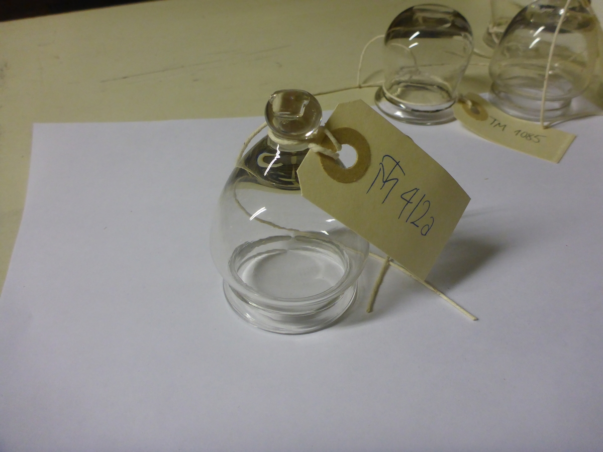 Koppeglass blåst i blankt glass. Formet som en klokke med en rund knapp øverst, og med glassmassen brettet ut som en brem nederst.