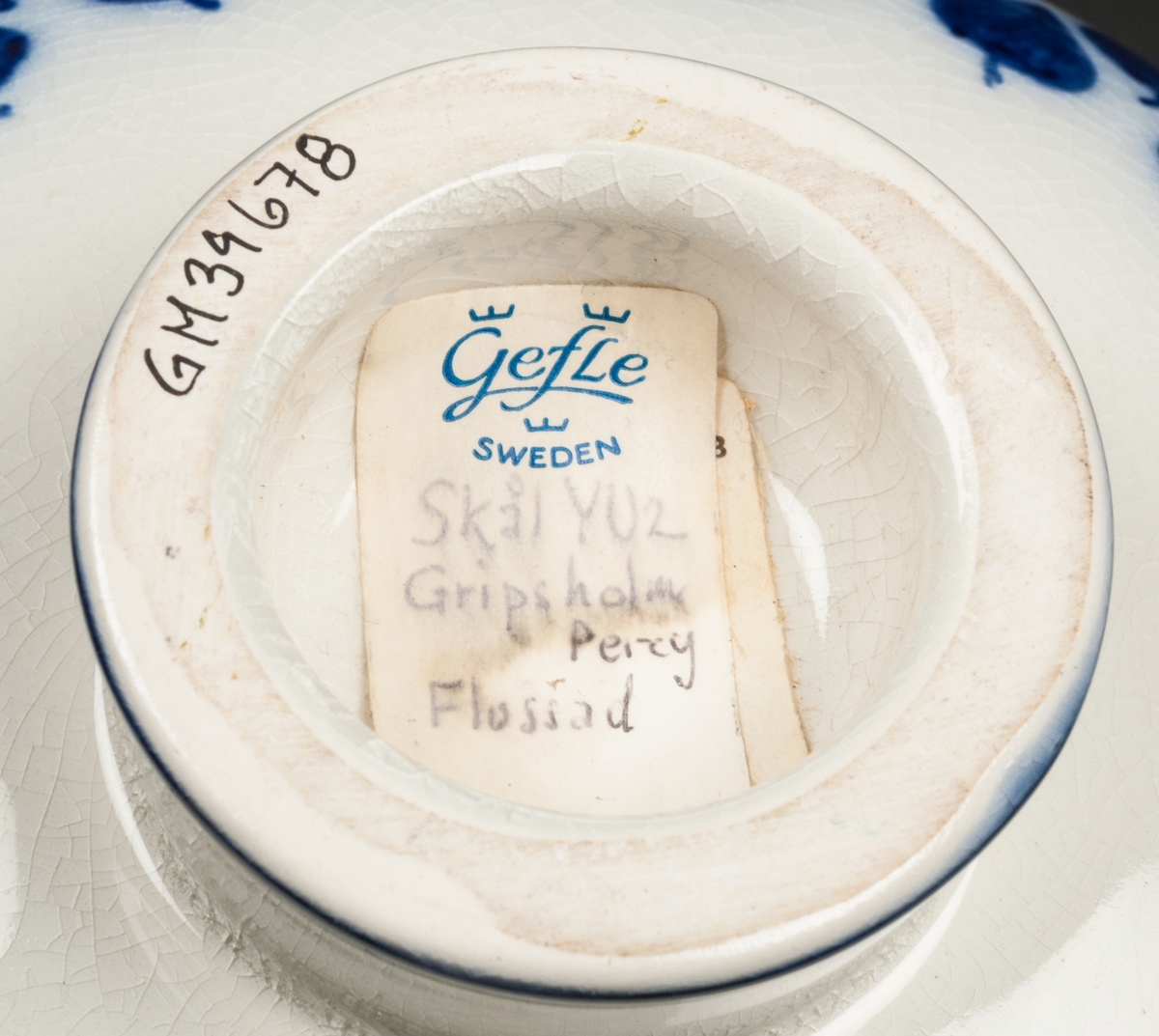 Skål på liten fot. Målade blå blad i glasyren, mot vit botten.
Etikett: Modell YU2. Gripsholm, Percy. Flossad.