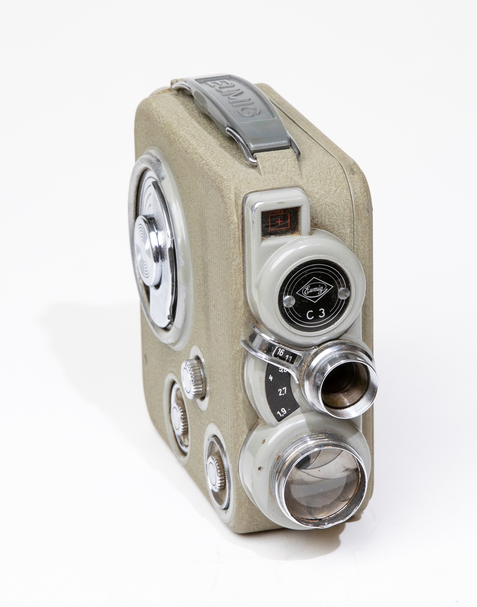 Filmkamera, dubbel-8 Eumig C 3, med kamerahus av grå metall, försedd med fjädermotor. Handtag av plast. Filmkameran är placerad i en kameraväska av brunt läder, JM 56584, och skruvas fast i väskan med en skruv. I kameraväskan förvaras ett litet linsskydd i en röd påse, och en fjärrutlösare.