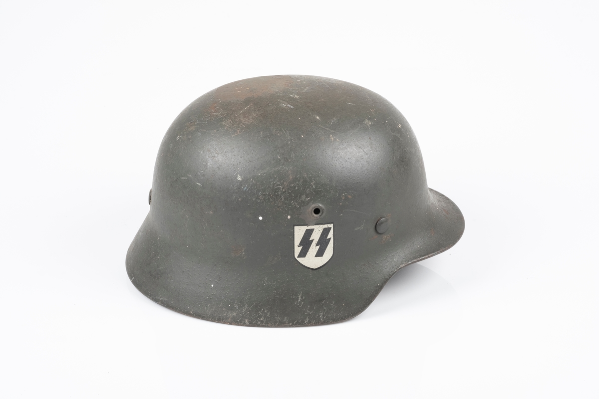 Tysk soldathjelm i metall med "SS" logo. Hjelmen har to sirkelrunde hull på hver side, som er symmetrisk plassert i forhold til hverandre. På innsiden er det festet en innerhjelm av lær med snor som brukes til å tilpasse hjelmen etter hodets størrelse. Hjelmen har lærrem med metallspenne. Hjelmen er malt brun-grønn.