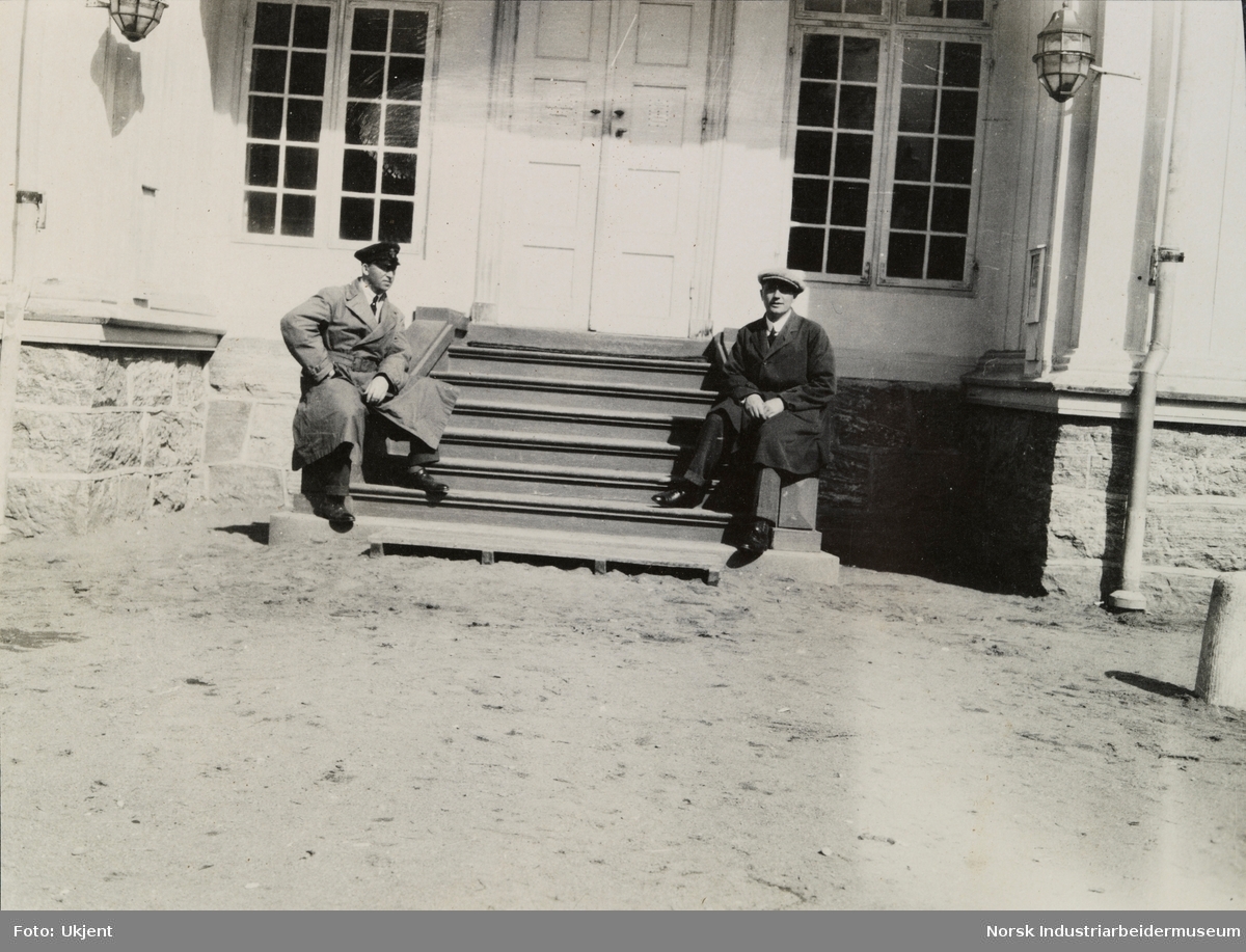 To menn sitter utendørs på hver sin side av trapp ved inngangsdør til antagelig Eidsvollbygningen