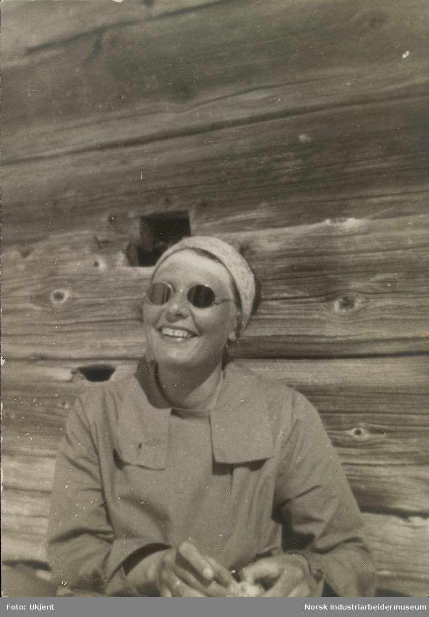 Portrett av smilende kvinne med pannebånd og runde solbriller mot yttervegg