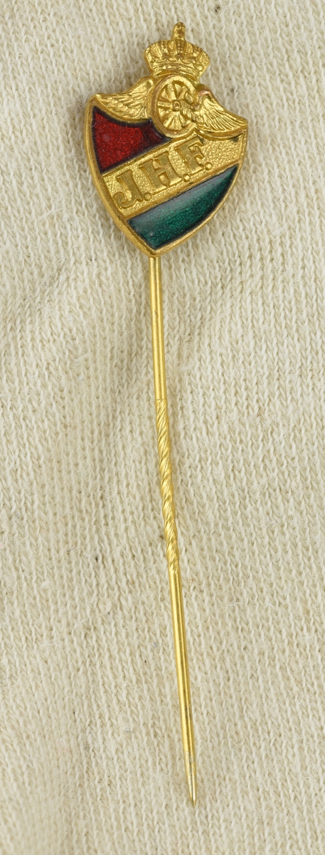 Kavajnål bestående av en platta i form av en sköld, krönt av ett bevingat hjul med krona, med en fastlödd nål på baksidan. Nålen är tillverkad av mässing med emalj. 
Skölden är indelat i tre fält i färgerna rött, gult och grönt.
